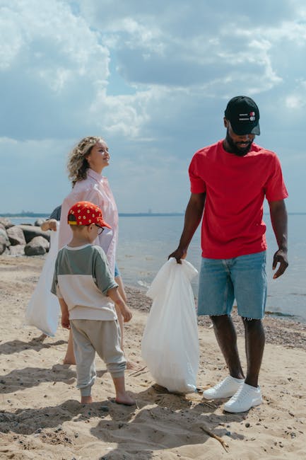 A man wearing a black cap, red t-shirt, and denim shorts is standing on a sandy beach, holding a large white plastic bag. Behind him, a woman with curly blonde hair dressed in a light pink jacket and trousers stands near the shoreline, along with a young boy wearing a red cap with white polka dots and grey and white clothing, also holding a white plastic bag. In the background, there are rocks along the water's edge, and the scene is set under a partly cloudy sky with blue and grey tones. The environment appears to be a natural coastal area, and the individuals seem to be participating in a beach clean-up activity, aligning with the context of rubbish removal services provided by Rubbish Removal Hampstead.