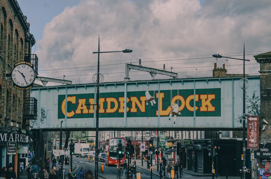 A busy urban street scene featuring a large overhead railway bridge with a prominent Camden Lock sign painted on its side in bold yellow and red letters. Below the bridge, there are multiple pedestrians walking along the pavement, some waiting at traffic lights, and a red double-decker bus moving through the intersection. On the left, part of a historic brick building with a clock displaying approximately 2:25 is visible, along with a street-level shopfront. Surrounding the scene are several tall street lamps, signage, and traffic management elements, all under an overcast sky with scattered clouds. The environment captures the vibrant atmosphere typical of Camden Town, with a mixture of modern transportation and urban architecture, illustrating the area's characteristic street life without directly referencing waste management or rubbish removal services.