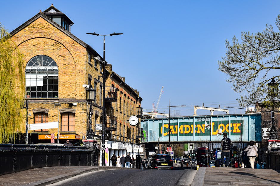 A street scene in Hampstead featuring a historic brick building with large arched windows and a small dormer window on the roof. The building's facade is constructed from yellowish-brown bricks with black accents around the windows and a black roof. In front of the building, there is a busy pavement area with pedestrians walking and standing, some carrying bags. To the right, a bridge with a painted sign reading 'Camden Lock' spans the street, indicating a nearby market or cultural area. Several black vehicles are parked along the curb and a black van is partially visible behind the bridge. A lamppost with two horizontal, modern black lights is positioned on the sidewalk, and a round clock is mounted on the building's exterior wall. The scene is illuminated by bright daylight, with a clear blue sky overhead. Leafless or sparsely leaved trees are visible on the right side of the image, providing some green and natural contrast to the urban environment. This street image highlights typical elements involved in private rubbish disposal and waste management services, as offered by companies like Rubbish Removal Hampstead, who may handle waste removal needs in such busy, historical areas.