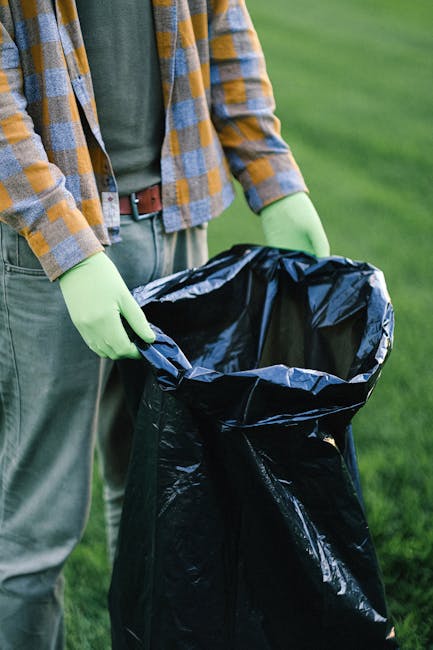 A person wearing a plaid shirt with yellow, blue, and gray tones and a dark green T-shirt underneath is holding open a large black plastic rubbish bag with lime green gloves. The individual is standing outdoors on a grassy area, with a blurred background of green lawn. The rubbish bag appears to be partially filled, ready for waste disposal, and the person’s stance suggests they are preparing to deposit rubbish into the bag. The scene emphasizes the act of waste collection or on-site rubbish clearance, which could relate to private waste handling services offered by companies such as Rubbish Removal Hampstead. The natural outdoor setting, along with the focus on the rubbish bag and gloves, highlights the practical aspect of rubbish collection and disposal, supporting efficient rubbish removal and environmentally responsible waste management practices.