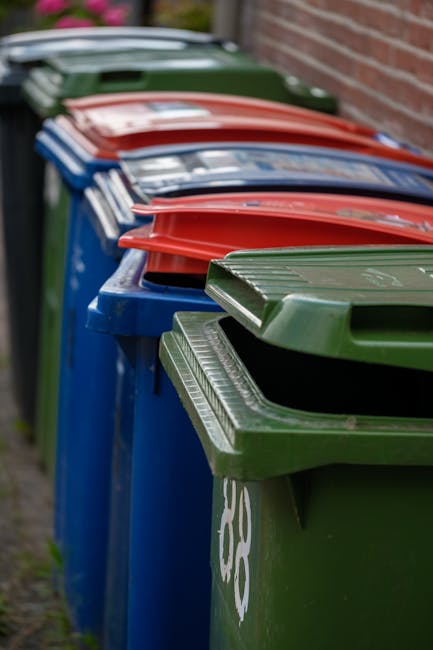 A row of large, brightly coloured wheelie bins lined up along a paved pathway next to a brick wall. The bins are primarily green, blue, red, and darker green, each with a rectangular lid that is slightly raised, showcasing textured patterns or smooth finishes. The bins are made of durable plastic material with a glossy surface, and some lids have subtle ridges or embossed designs. They are positioned close together, slightly angled, with the green bin in front displaying a white recycling symbol. The background includes a blurred view of the pavement and part of the brick wall, suggesting an outdoor refuse collection area near a residential or commercial building. The scene is evenly lit with natural daylight, emphasizing the vivid colours of the bins and the clean, orderly arrangement suitable for waste management or alternative rubbish removal services like those offered by Rubbish Removal Hampstead, emphasizing efficient on-site or private waste handling options.