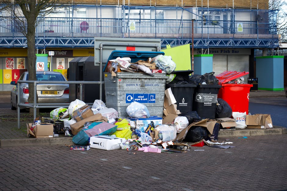 A large collection of mixed waste, including cardboard boxes, plastic bags, paper, and packaging materials, is piled around and inside open-top bins on a paved urban street. The waste is overflowing from the bins, with some items spilling onto the ground, and appears to include various household and office rubbish such as flattened boxes, crumpled paper, and plastic wrapping. The bins occupy the foreground, with a street railing separating them from parked cars behind, including a silver vehicle with a partially visible license plate. In the background, there is a commercial building with storefronts and signage, and the upper levels are covered with blue scaffolding or safety netting, indicating ongoing construction or maintenance work. The scene is lit with natural daylight, highlighting the disorganized state of the waste collection area. This image illustrates the need for proper rubbish management services, like those offered by Rubbish Removal Hampstead, to handle excess waste from businesses and help maintain a clean environment in urban areas.
