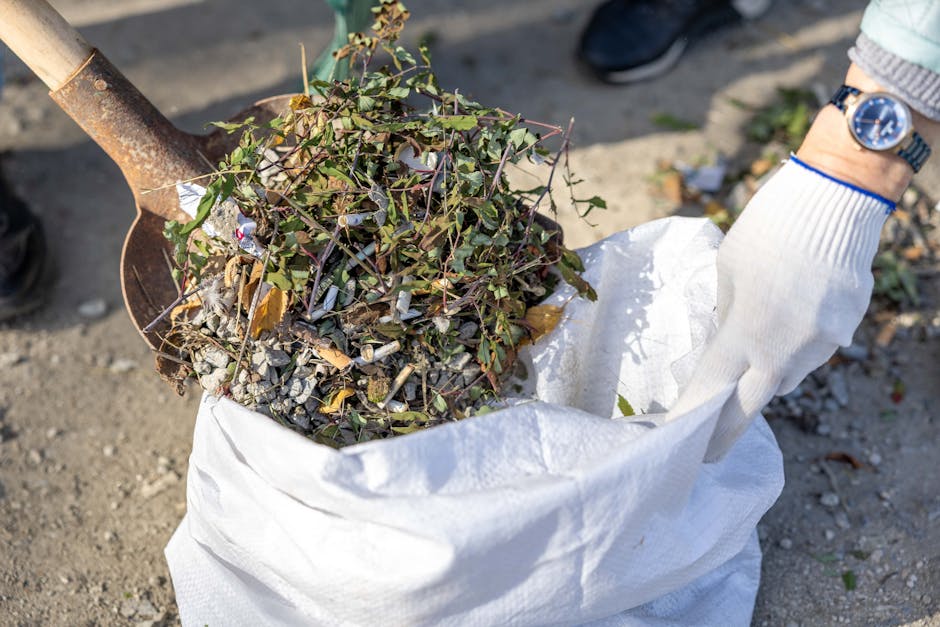 A close-up view of a person's hand wearing a white glove, holding a large white heavy-duty rubbish bag filled with mixed garden waste, including small branches, leaves, and soil-covered debris. A rusty spade with a wooden handle is partially inserted into the bag, suggesting ongoing collection or transfer of waste. The scene takes place outdoors on a gravel or dirt surface, with the person's wristwatch visible on their wrist. The natural lighting highlights the varied textures of the leaves, twigs, and soil, embodying the practical process of waste clearance commonly handled by services such as Rubbish Removal Hampstead. The image captures a moment of private waste handling, exemplifying an alternative to municipal rubbish collection and emphasizing efficient, on-site rubbish removal.