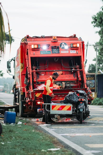 A sanitation worker in a red and yellow uniform is collecting waste near the rear of a large red rubbish collection truck parked beside a paved road. The worker is focused on loading debris, with the back of the truck open, revealing the empty hopper used for waste disposal. The truck's exterior has reflective yellow and red safety markings, and small lights on the upper section. A black garbage bag rests on a small metal cart attached to a motorbike parked nearby. In the background, there are trees, utility poles, and a signboard, suggesting an urban or suburban environment. The scene is illuminated by natural daylight beneath a cloudy sky, and some litter can be seen on the ground, indicating waste collection activity typical of private or municipal rubbish removal services. The image visually aligns with waste management operations, such as those provided by companies like Rubbish Removal Hampstead, that handle on-site clearance and independent waste collection.