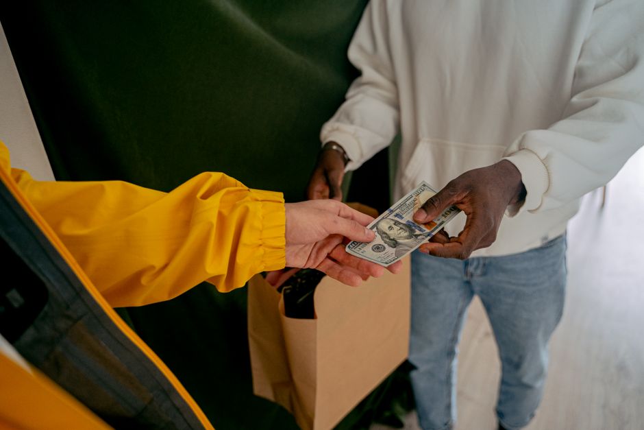 A person wearing a yellow waterproof jacket is receiving a stack of US hundred-dollar bills from another individual dressed in a white hoodie and light blue jeans. The exchange occurs near a green upholstered seat, with the person in the hoodie holding the cash with both hands. In the background, a brown paper bag rests on the floor, suggesting a setting related to waste disposal or collection. The scene is indoors, with natural lighting highlighting the textures of the paper money and fabric clothing, and the overall neutral tone emphasizes a straightforward transaction. This image subtly reflects forms of private waste handling, such as cash payments for rubbish removal services, aligning with the broader context of independent collection methods distinct from local authority rubbish collection in Hampstead, as discussed on the Rubbish Removal Hampstead website.